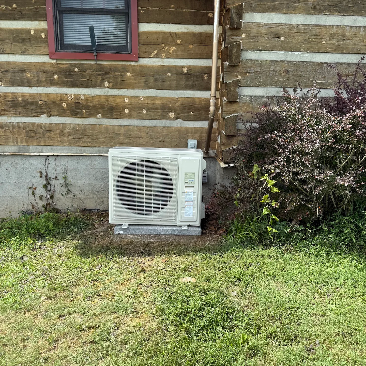 A ductless mini split outdoor unit set beside the log wall of a log home at dusk