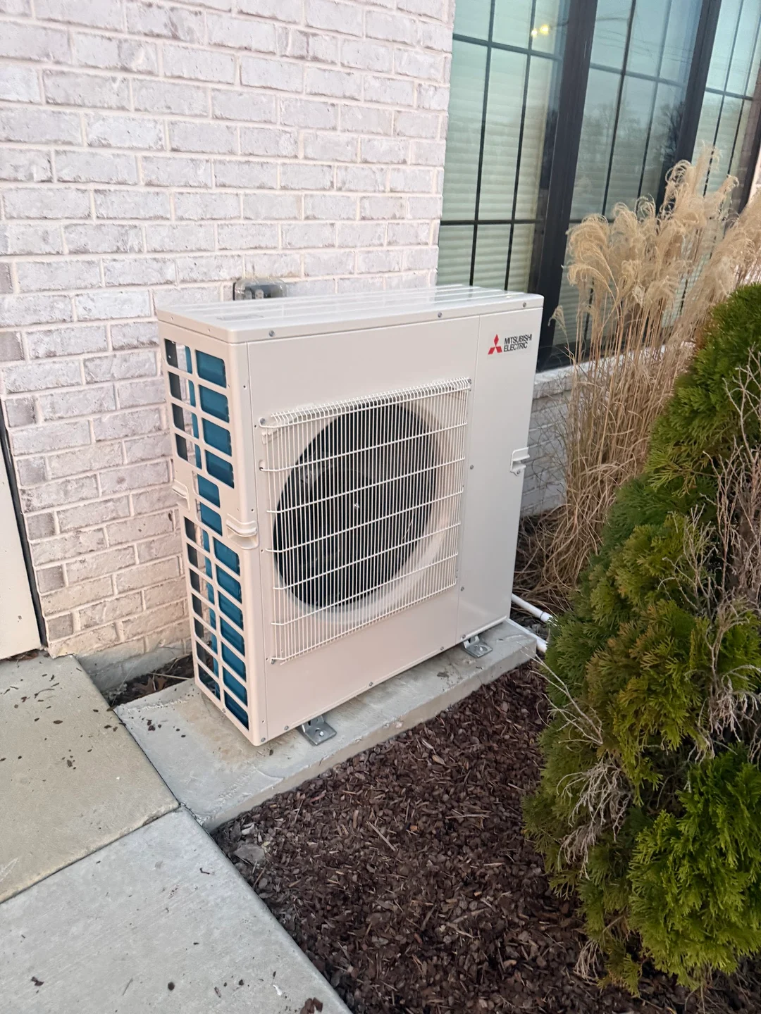 A Mitsubishi Electric multi-zone heat pump outdoor unit on a pad against a white-painted brick wall